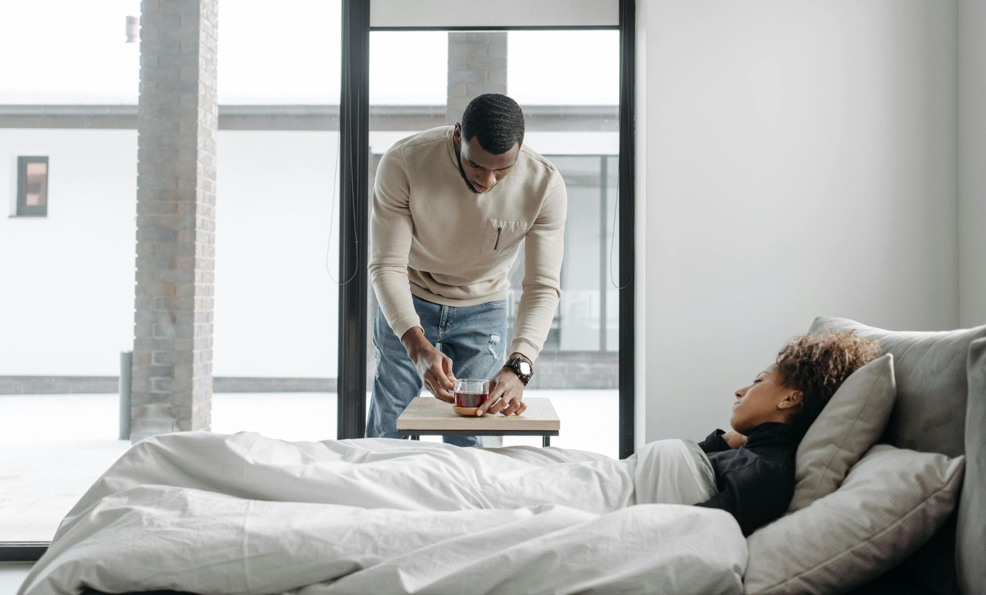 A man cares for his sick wife in bed, bringing her a cup of tea indoors.